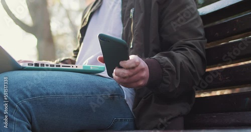 Close up of young man working on laptop in park on bench. Fall day. Outdoors. Male freelancer with computer at nature. Checking mobile phone. guy using smartphone and device remote. photo