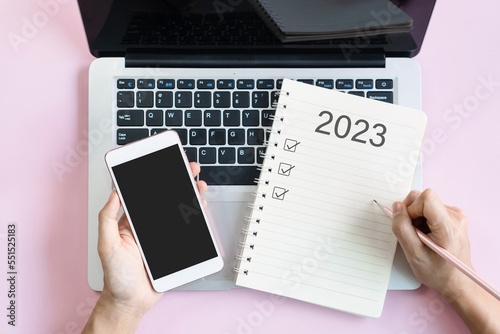 Flat lay of woman hand writing 2023 to do list on notebook while holding mobile phone with blank screen. Copy space, top view. 2023 planning concept. photo