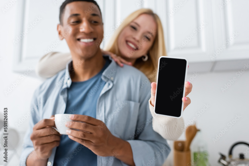 happy blonde woman holding smartphone with blank screen near african american man with coffee cup on blurred background