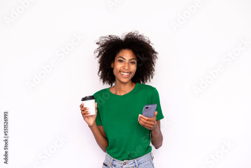 Portrait of happy young woman holding mobile phone and coffee over white background. African American lady wearing green T-shirt and jeans looking at camera and smiling. Mobile technology concept