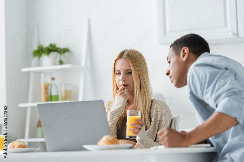 thoughtful woman with orange juice looking at laptop near blurred african american boyfriend in kitchen thoughtful woman with orange juice looking at laptop near blurred african american boyfriend in kitchen