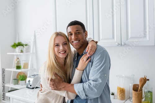 cheerful interracial couple looking at camera while embracing in kitchen