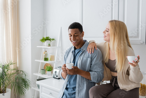 joyful blonde woman sitting on kitchen worktop near african american boyfriend with coffee cup