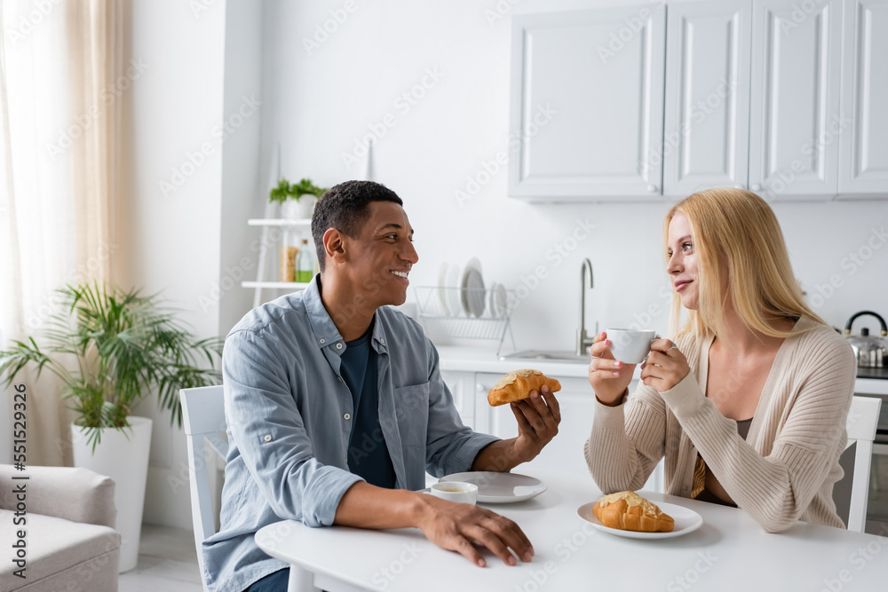 happy interracial couple looking at each other while having breakfast with coffee and delicious croissants
