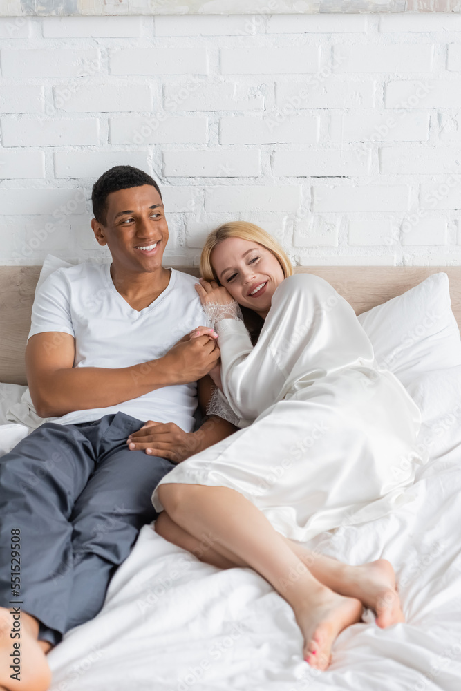 happy african american man holding hand of blonde girlfriend in white robe lying on bed in morning happy african american man holding hand of blonde girlfriend in white robe lying on bed in morning