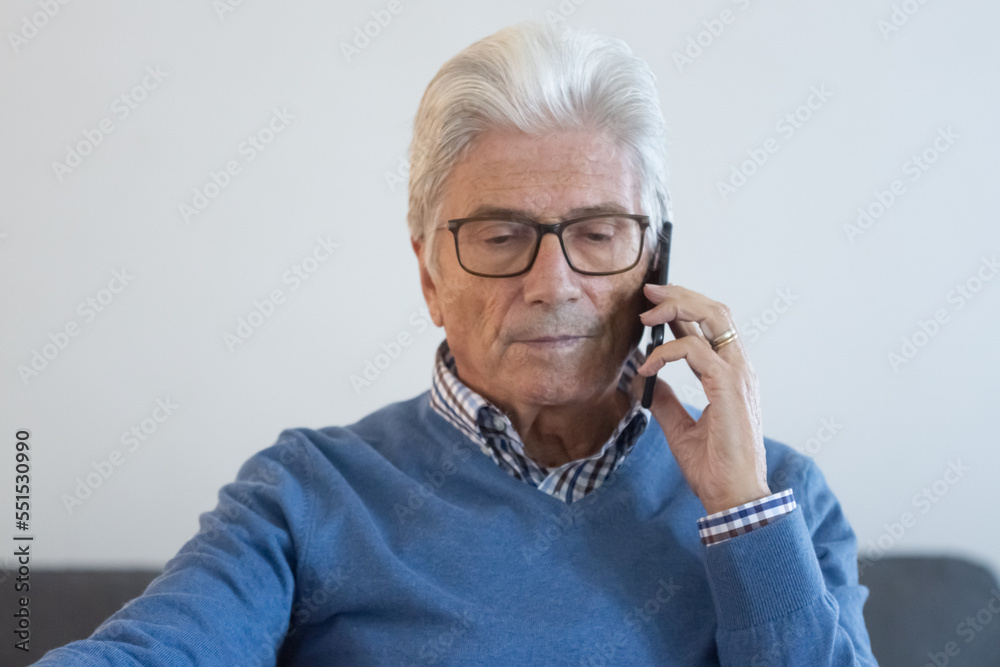 Serious senior man talking on phone in living room. Front view of mature businessman having conversation with colleague, sitting at table in home office. .Digital devices, communication concept Serious senior man talking on phone in living room. Front view of mature businessman having conversation with colleague, sitting at table in home office. .Digital devices, communication concept