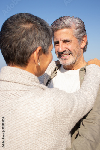 Happy senior couple spending time at seashore on sunny day. Smiling bearded man hugging his wife while couple looking at each other. Over the shoulder shot. Romance, leisure concept