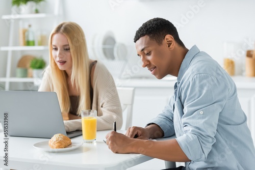 african american man writing in notebook and blonde woman working on laptop near breakfast in kitchen