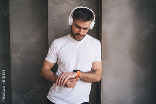 Waiting man listening to music in headphones and standing near gray wall in gym