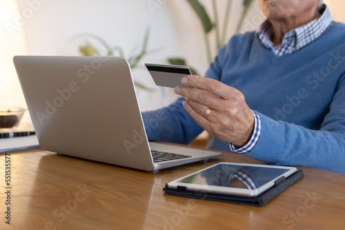 Elderly man using laptop for shopping online at home. Unrecognizable mature man sitting at table in living room, buying services through internet, paying with credit card. Online shopping concept