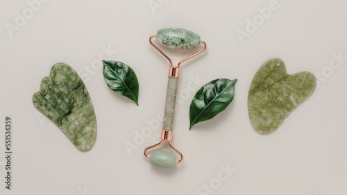 Jade face roller and gua sha stone massager with green leaves on pastel background, banner size. Spa face and body care tool. Gua sha stone scraper, natural skincare