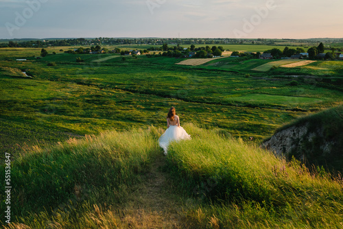 Bride holding her wedding dress and running in a field on the road with sunlight. Woman in the mountains top at sunset. Girl walking on meadow in green grass. Back view.