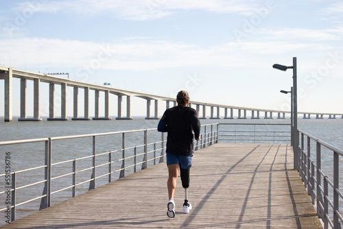 Back view of man with mechanical leg running on embankment. Caucasian man in sportive clothes jogging on summer day. Sport, leisure, disability concept photo