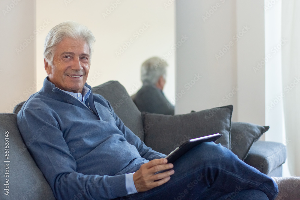 Smiling handsome senior man sitting on sofa and looking at camera. Positive successful aged businessman using tablet at home. Comfort concept