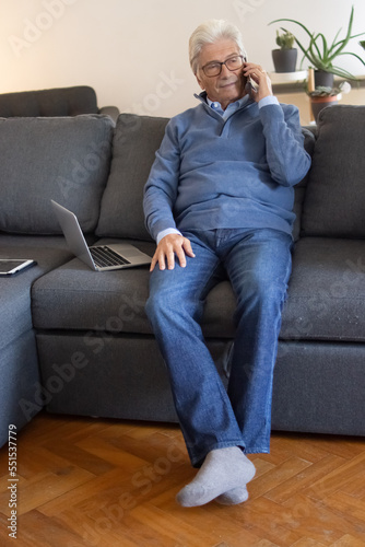 Busy aged man calling by cellphone. Content senior businessman in casual outfit sitting on sofa with laptop and tablet at home. Remote work concept