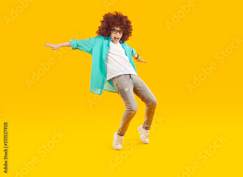 Happy kid dancing and having fun. Funny cheerful excited child boy in casual clothes and curly wig dancing on an amber orange yellow color studio background