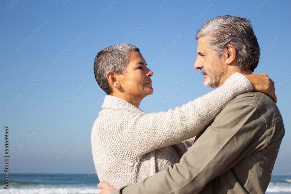 Gray-haired senior couple hugging at seashore on sunny autumn day. Short-haired middle-aged lady and bearded senior man looking at each other. Side view. Relationship, love, traveling concept Gray-haired senior couple hugging at seashore on sunny autumn day. Short-haired middle-aged lady and bearded senior man looking at each other. Side view. Relationship, love, traveling concept
