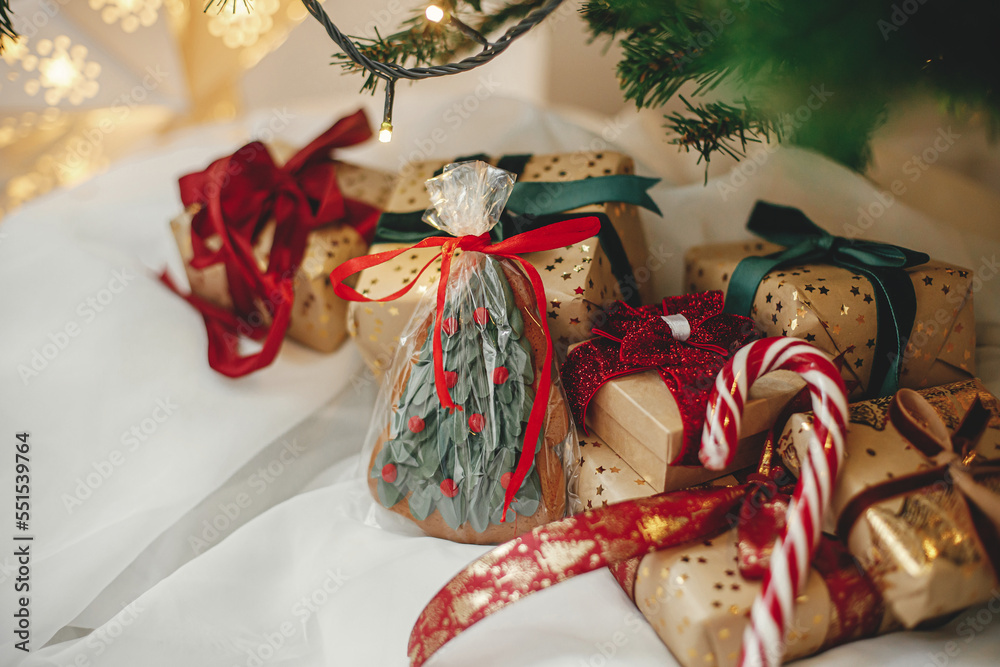 Merry Christmas! Stylish christmas gifts with cookie and candy cane under christmas tree with golden lights. Wrapped christmas presents in golden paper with red and green ribbons in festive room