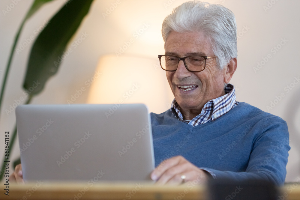 Happy senior man having video call with friends at home. Smiling mature man in eyeglasses sitting at table in living room, looking at screen and talking. Greeting, digital devices concept Happy senior man having video call with friends at home. Smiling mature man in eyeglasses sitting at table in living room, looking at screen and talking. Greeting, digital devices concept