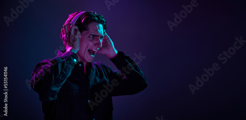 Portrait of young man in casual black shirt isolated over gradient dark purple background in neon light. Music in headphones. Concept of human emotions, facial expression.