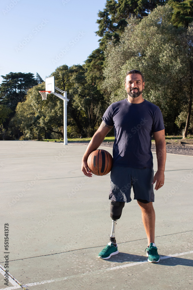 Smiling Hispanic man with physical disability standing at basketball court in park on sunny day. Strong man with leg prosthesis doing sports outside. Amputee sport, motivation concept Smiling Hispanic man with physical disability standing at basketball court in park on sunny day. Strong man with leg prosthesis doing sports outside. Amputee sport, motivation concept