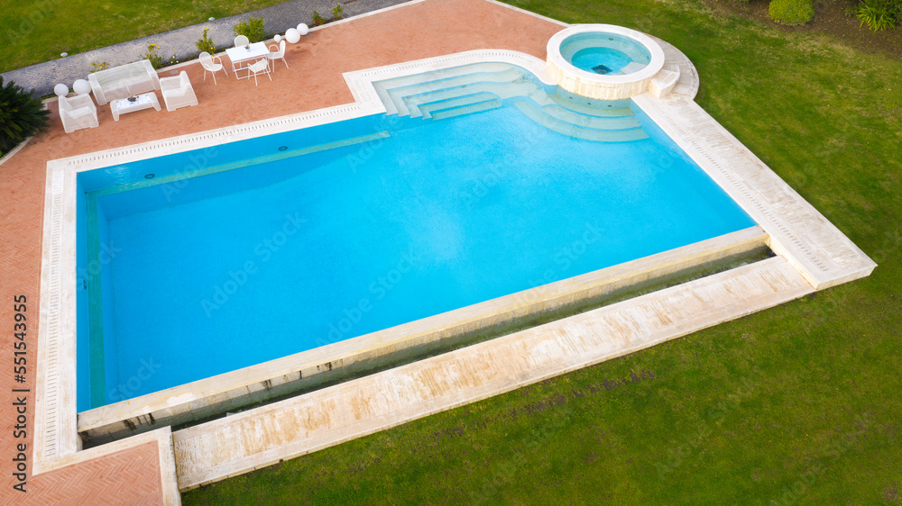 Aerial view of a blue pool with stairs to descend and climb into the water. Around marble tiles. There are empty loungers by the pool.