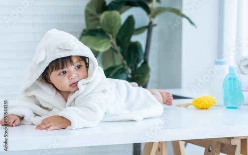 Adorable caucasian little baby daughter girl, wearing cute white bathrobe, taking bath in morning, sitting in baby bathtub, laughing, smiling with happiness at home or house. Kid Development Concept.