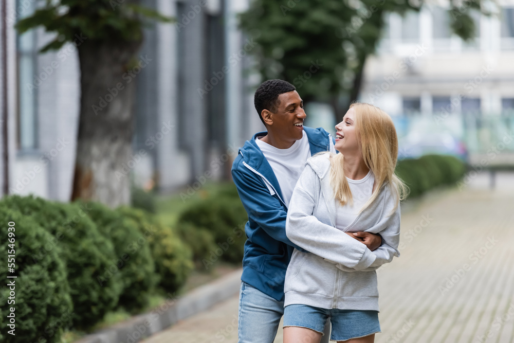 joyful african american man embracing young blonde girlfriend on blurred street in city