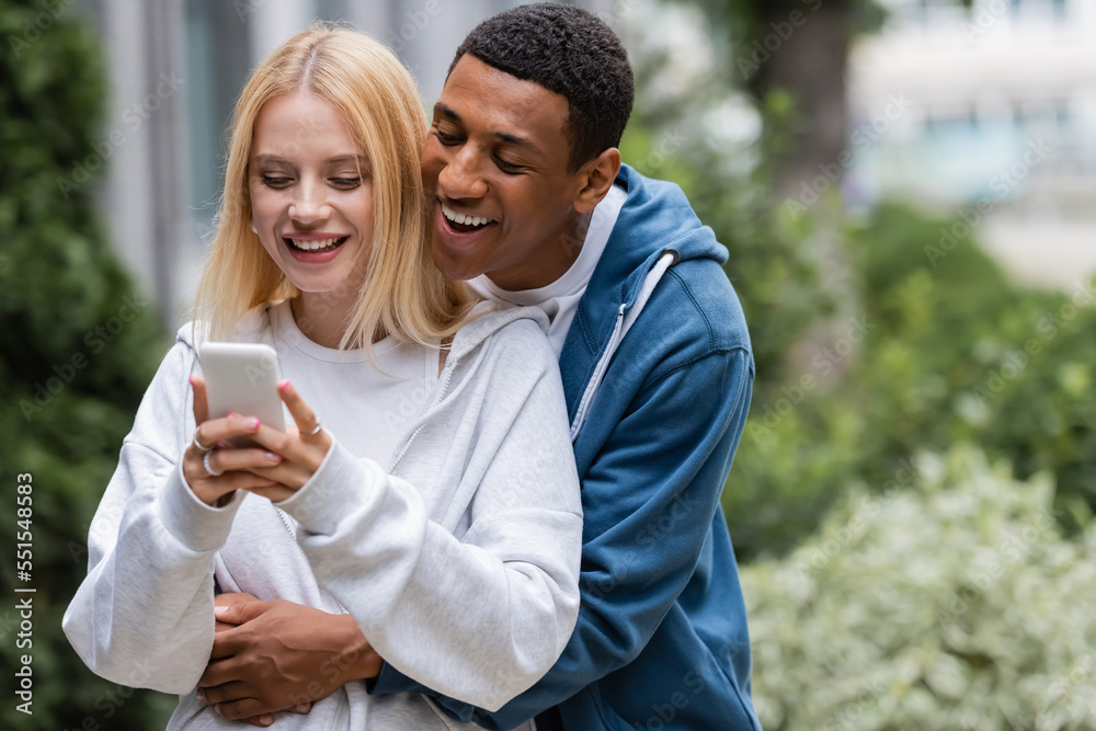smiling african american man embracing blonde girlfriend messaging on smartphone outdoors smiling african american man embracing blonde girlfriend messaging on smartphone outdoors