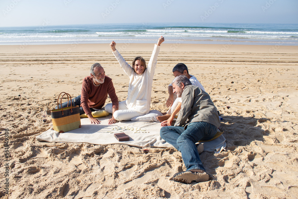 Senior friends playing dominoes outdoors on sunny day while enjoying picnic together at the beach. Middle-aged blonde woman raising arms in triumph having won the game. Entertainment, leisure concept