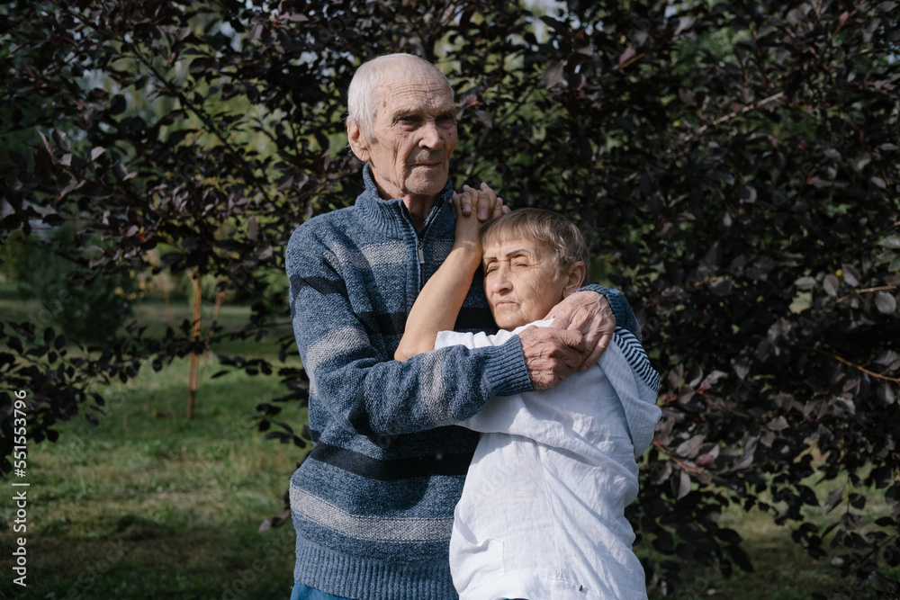 Fototapeta premium Elderly couple husband and wife standing outdoor in summer park, hugging relaxing enjoying sunny day and love