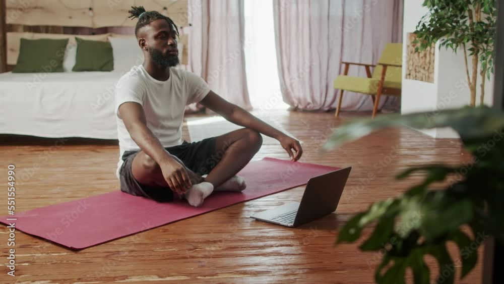 Young Beginer African American man in sportswear practicing yoga at home. Male sit on mat in lotus position on red fitness mat in living room in front of open laptop, healthcare and exercise mental