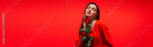 Trendy brunette model in jacket holding rose and closing eyes isolated on red, banner