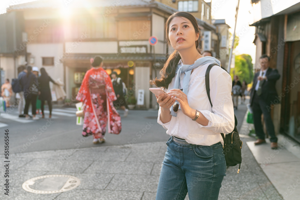 backlight shot of asian Japanese girl tourist looking into distance with curiosity while using navigation app on phone at Ninenzaka and Sannenzaka in Kyoto japan with a kimono girl at background