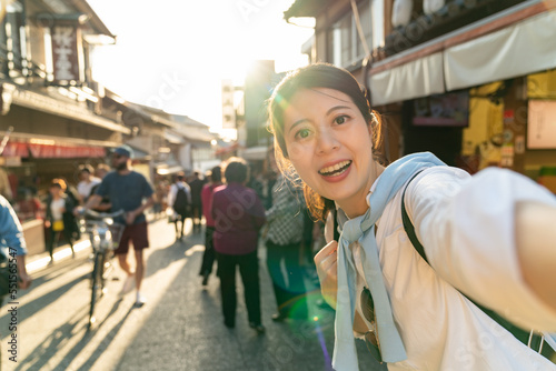 smiling asian Japanese girl visitor looking at camera while taking selfie picture at retro shopping street of Ninenzaka and Sannenzaka in backlight at sunset in Kyoto japan photo