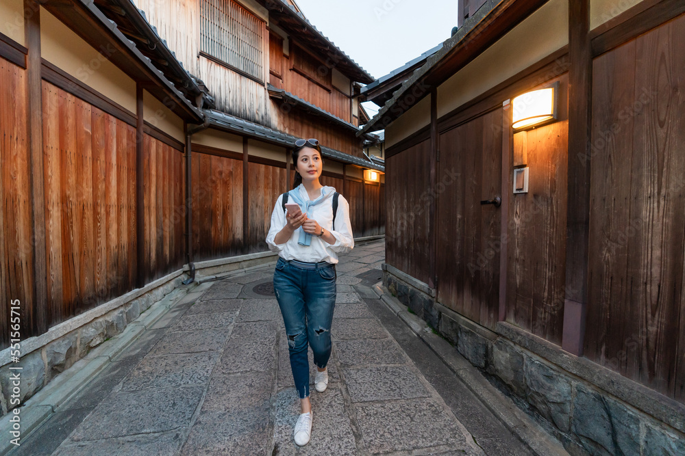 full length of leisure asian Japanese woman backpacker using gps app on mobile phone while walking through Ishibeikoji alley in gion Kyoto japan in the evening.
