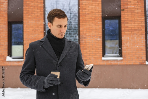 Serious adult businessman in winter coat surfing smartphone and having coffee break in city street against building 