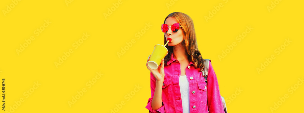 Portrait of stylish young woman drinking juice wearing pink jacket, sunglasses on yellow background