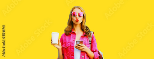 Portrait of stylish young woman with smartphone drinking coffee wearing pink jacket, sunglasses on yellow background