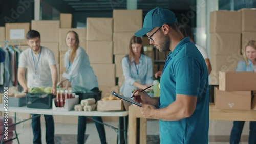 Male Volunteer Writing on Clipboard While Working in Food Bank Warehouse. Young Man Doing Inventory of Donated Goods in Volunteer Center While Group of Volunteers Sorting Humanitarian Aid