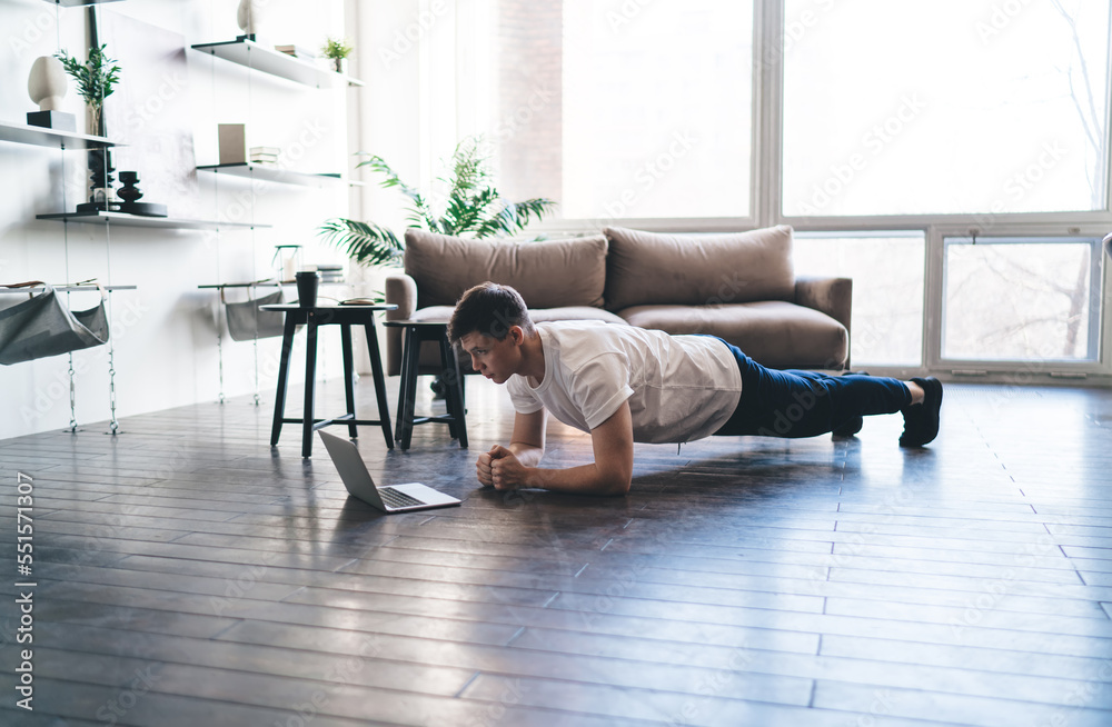 Concentrated man doing forearm plank exercise while watching sport lesson on laptop Concentrated man doing forearm plank exercise while watching sport lesson on laptop