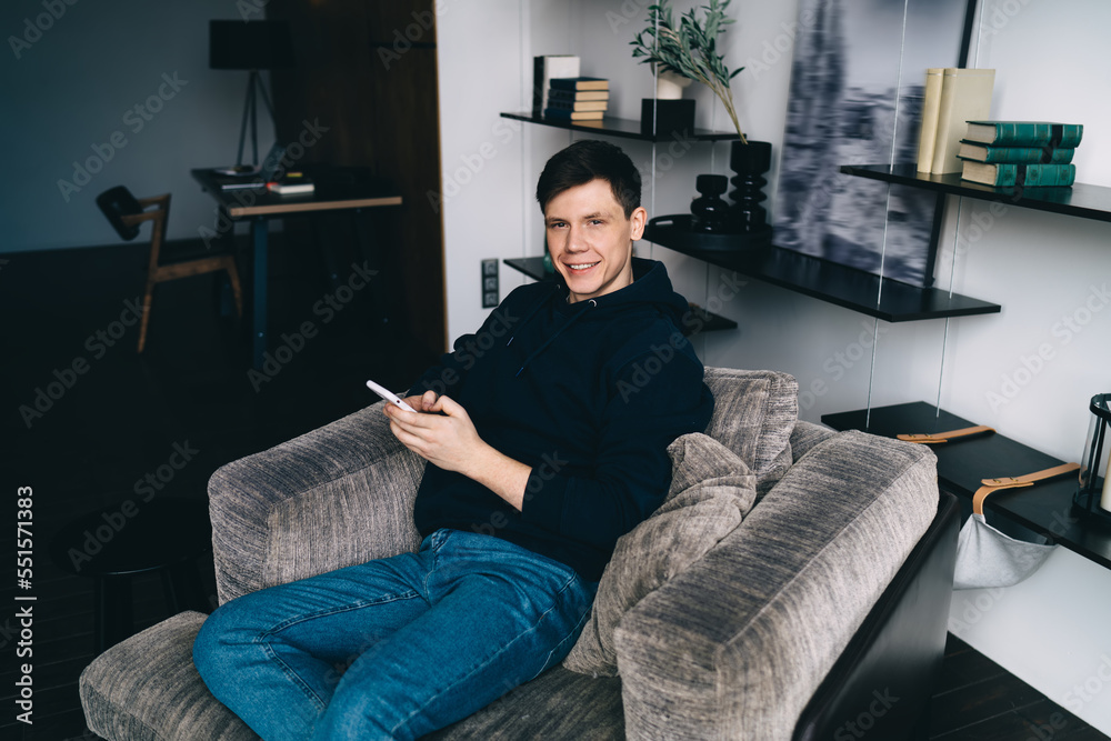 Smiling young man sitting on sofa with smartphone in hand in living room