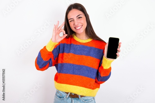 Young caucasian woman wearing colorful knitted sweater over white background holding in hands cell showing ok-sign