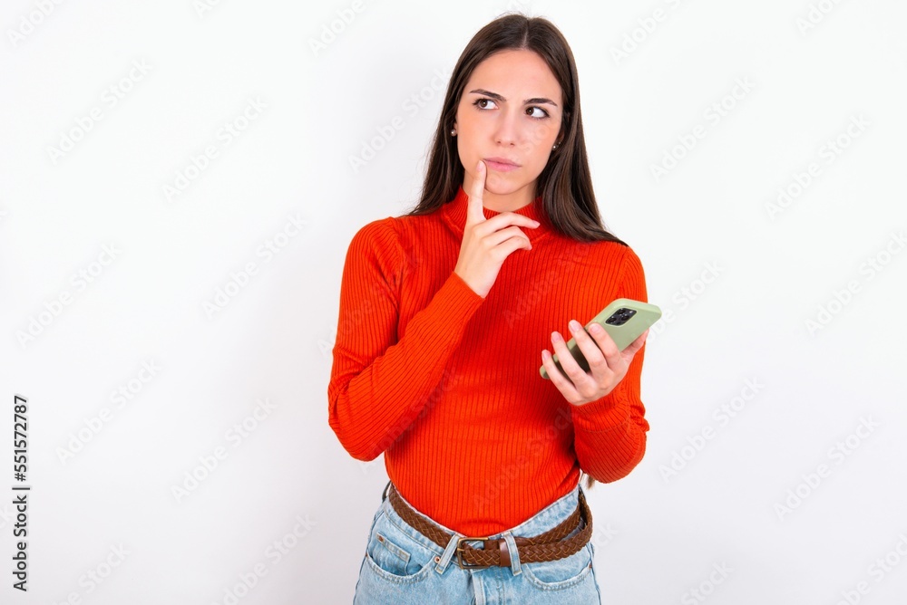 Young caucasian woman wearing red sweater over white background thinks deeply about something, uses modern mobile phone, tries to made up good message, keeps index finger near lips. Young caucasian woman wearing red sweater over white background thinks deeply about something, uses modern mobile phone, tries to made up good message, keeps index finger near lips.