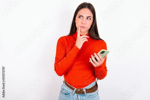 Young caucasian woman wearing red sweater over white background thinks deeply about something, uses modern mobile phone, tries to made up good message, keeps index finger near lips.