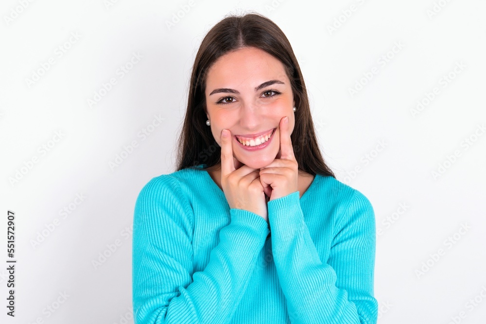 Happy Young caucasian woman wearing blue sweater over white background with toothy smile, keeps index fingers near mouth, fingers pointing and forcing cheerful smile Happy Young caucasian woman wearing blue sweater over white background with toothy smile, keeps index fingers near mouth, fingers pointing and forcing cheerful smile