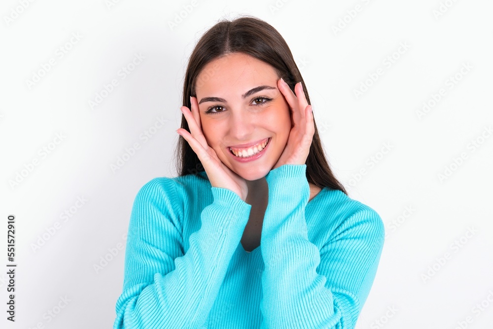 Happy Young caucasian woman wearing blue sweater over white background touches both cheeks gently, has tender smile, shows white teeth, gazes positively straightly at camera, Happy Young caucasian woman wearing blue sweater over white background touches both cheeks gently, has tender smile, shows white teeth, gazes positively straightly at camera,