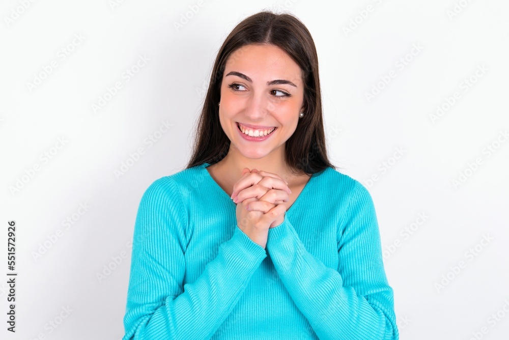 Happy Young caucasian woman wearing blue sweater over white background anticipates something awesome happen, looks happily aside, keeps hands together near face, has glad expression. Happy Young caucasian woman wearing blue sweater over white background anticipates something awesome happen, looks happily aside, keeps hands together near face, has glad expression.