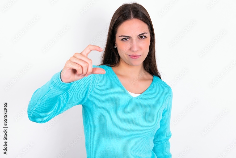 Displeased Young caucasian woman wearing blue sweater over white background shapes little hand sign demonstrates something not very big. Body language concept. Displeased Young caucasian woman wearing blue sweater over white background shapes little hand sign demonstrates something not very big. Body language concept.