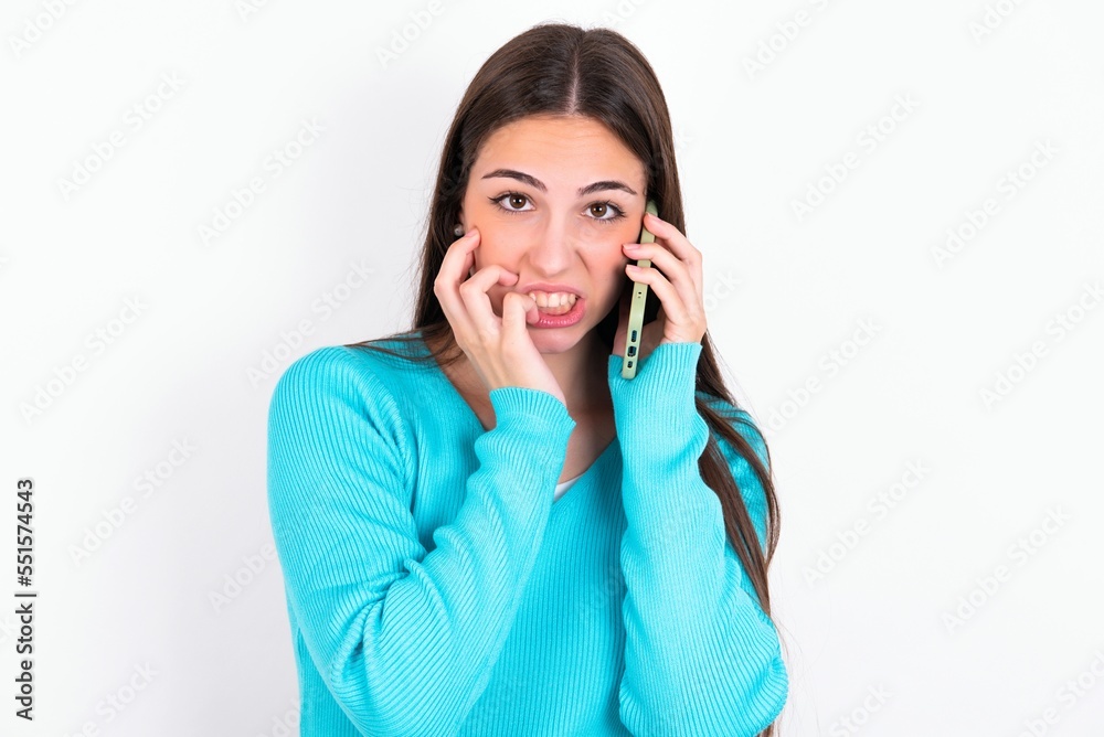 Afraid funny Young caucasian woman wearing blue sweater over white background holding telephone and bitting nails Afraid funny Young caucasian woman wearing blue sweater over white background holding telephone and bitting nails